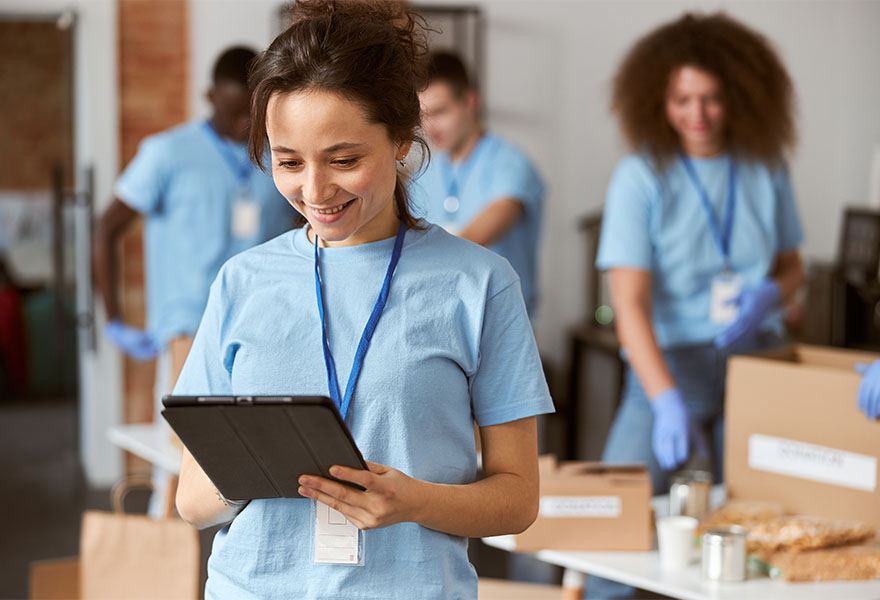 Portrait of charming young female volunteer in blue uniform using tablet pc and smiling while standing indoors. Team sorting, packing items in the background. Charitable foundation concept